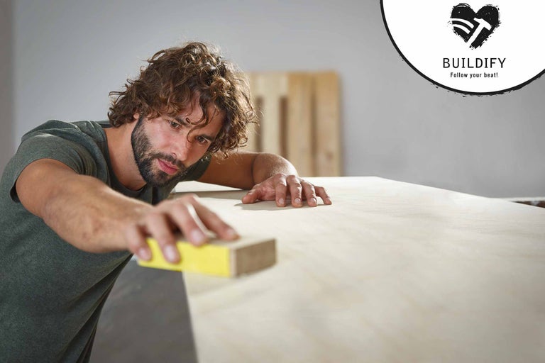 Ein Mann schleift Holz mit einem Schleifklotz. Buildify Logo.