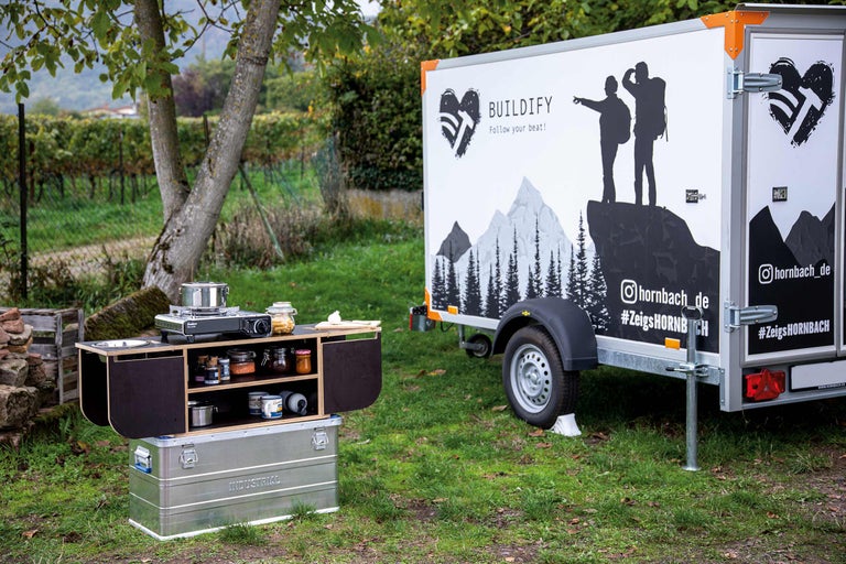Campingküche mit Kocher, Vorräten und Stauraum auf einem Feld mit Anhänger im Hintergrund.