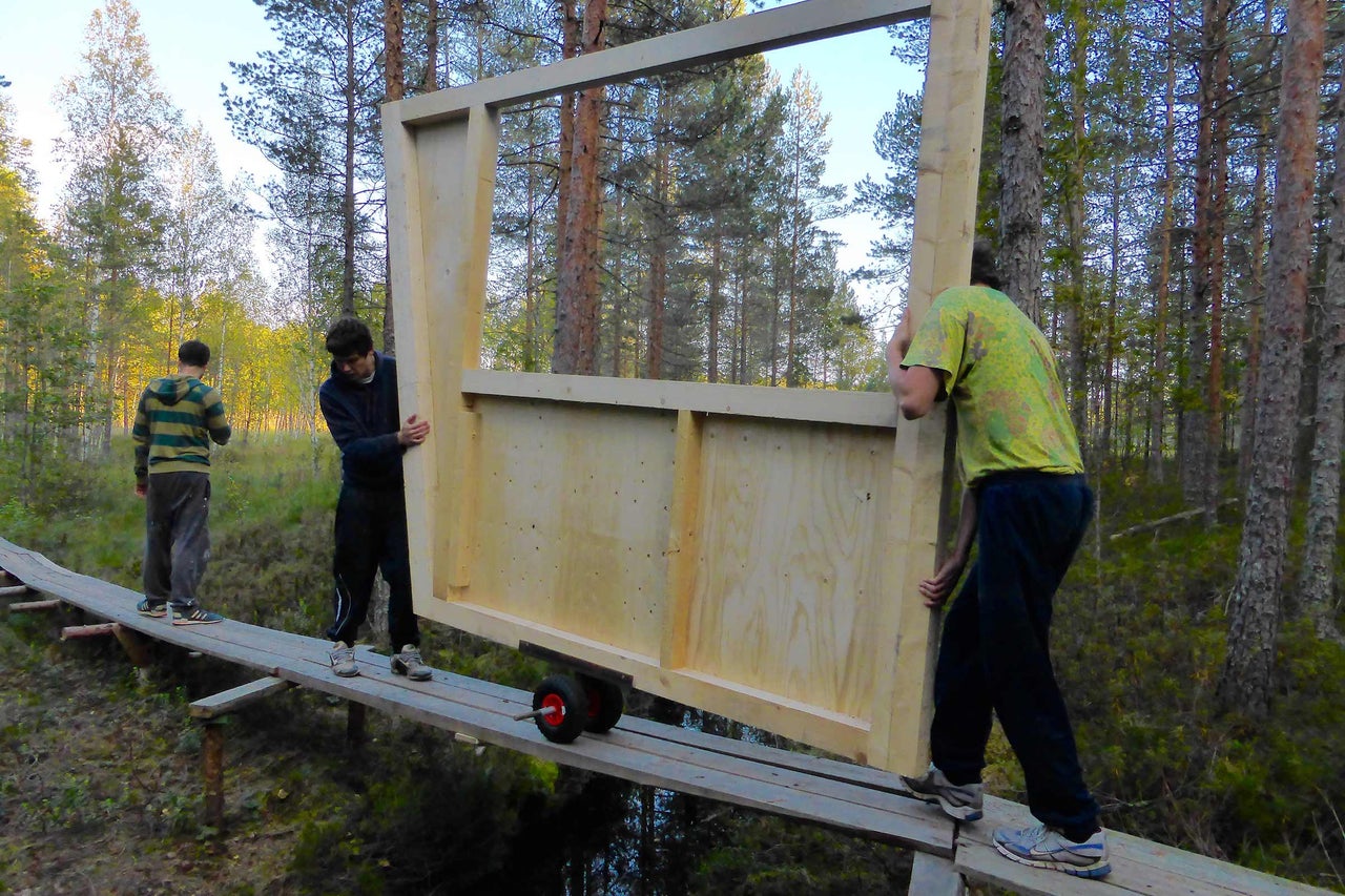 Zwei Personen transportieren ein Holzrahmenbauelement mit Rädern über eine Holzbrücke.