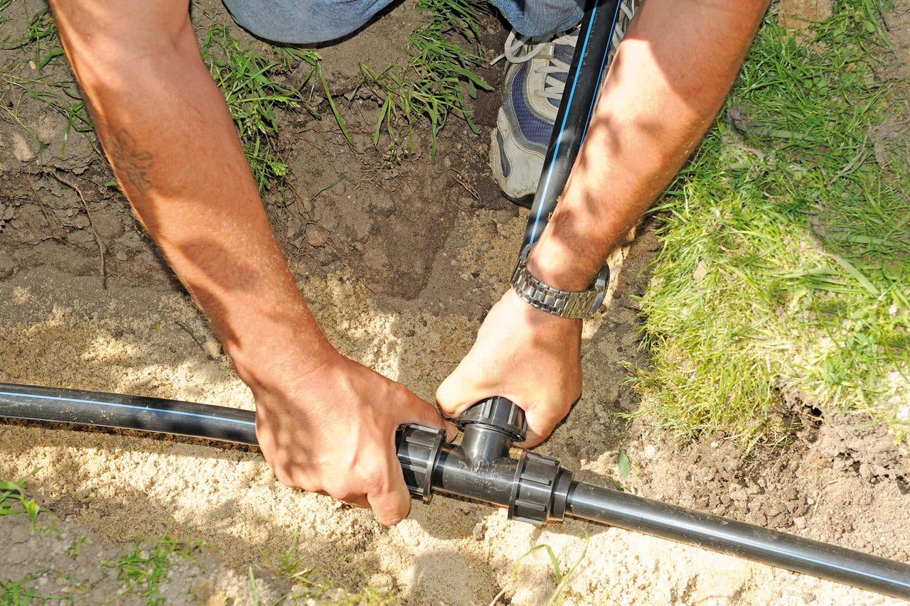 Person montiert ein T-Stück an einem schwarzen Bewässerungsrohr im Garten.