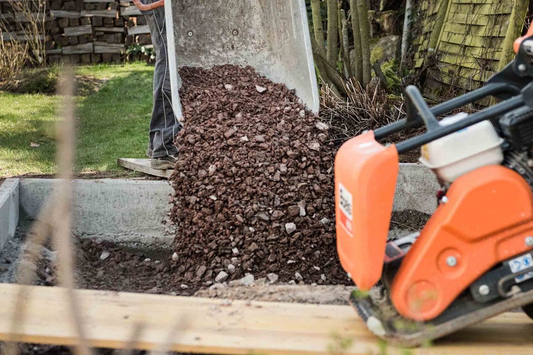 Eine Schubkarre wird in eine Baugrube entleert, während eine Rüttelplatte bereitsteht