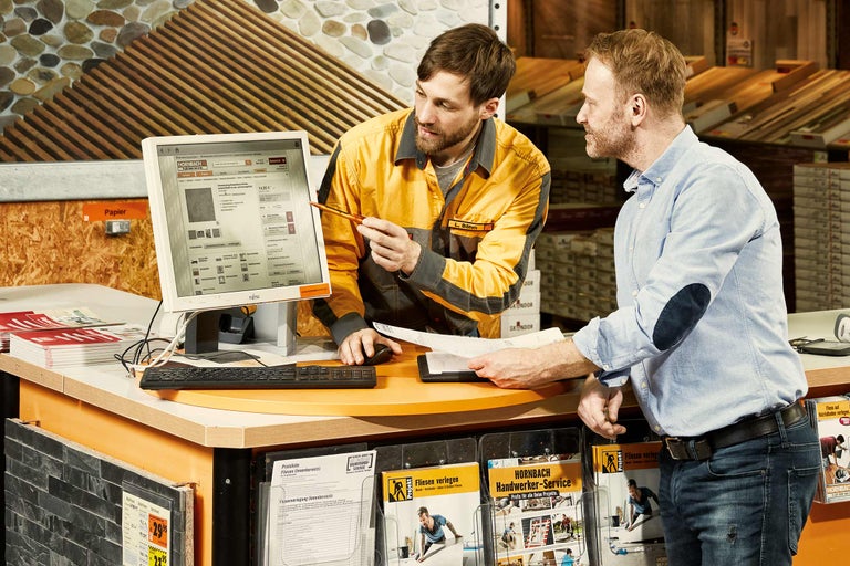 Zwei Mitarbeiter beraten sich an einem Beratungstisch bei Hornbach, auf dem ein Computer mit der Hornbach Webseite steht.