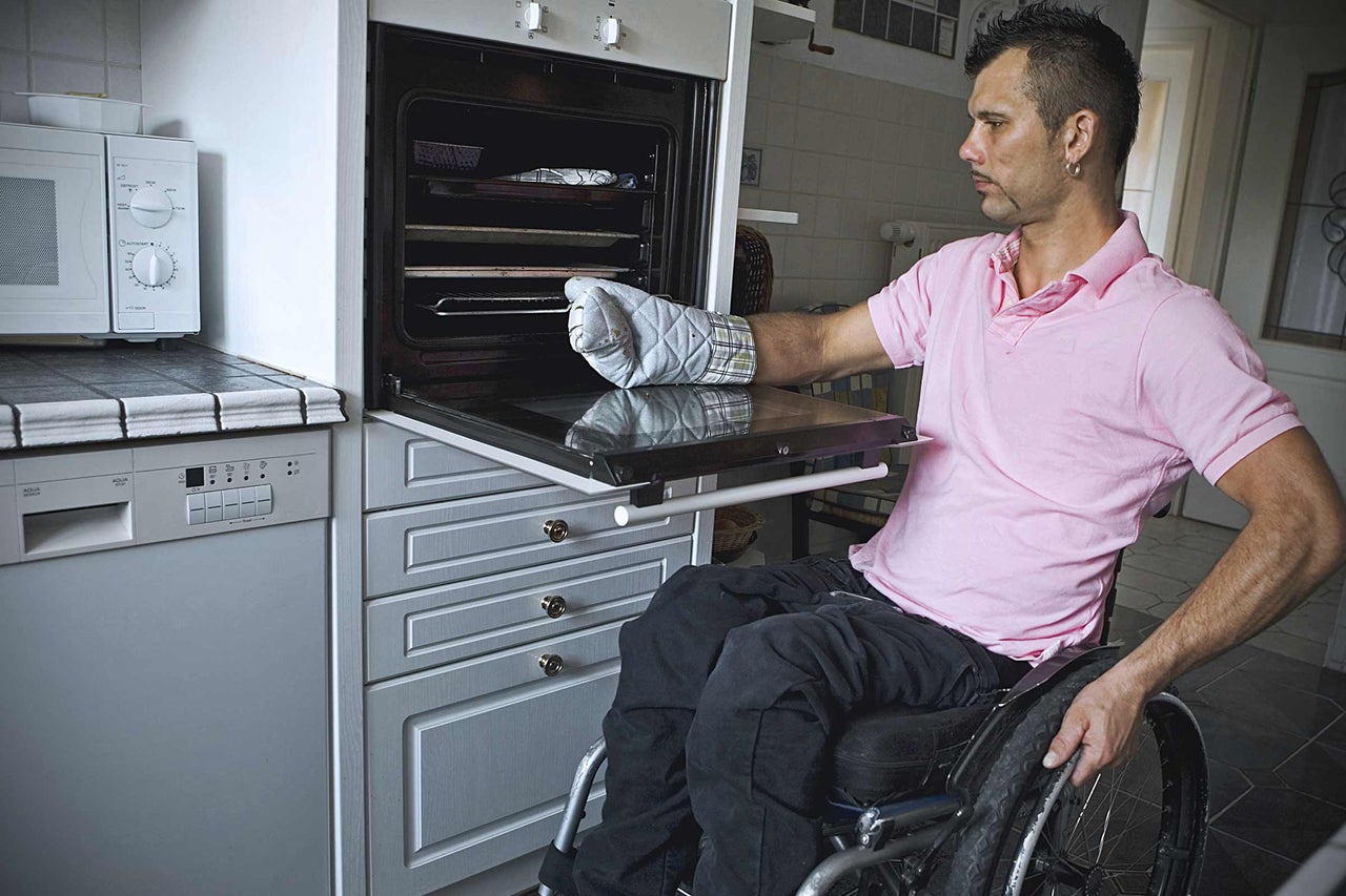 Mann im Rollstuhl benutzt einen Ofen mit Ofenhandschuh in einer Küche mit Mikrowelle und Geschirrspüler