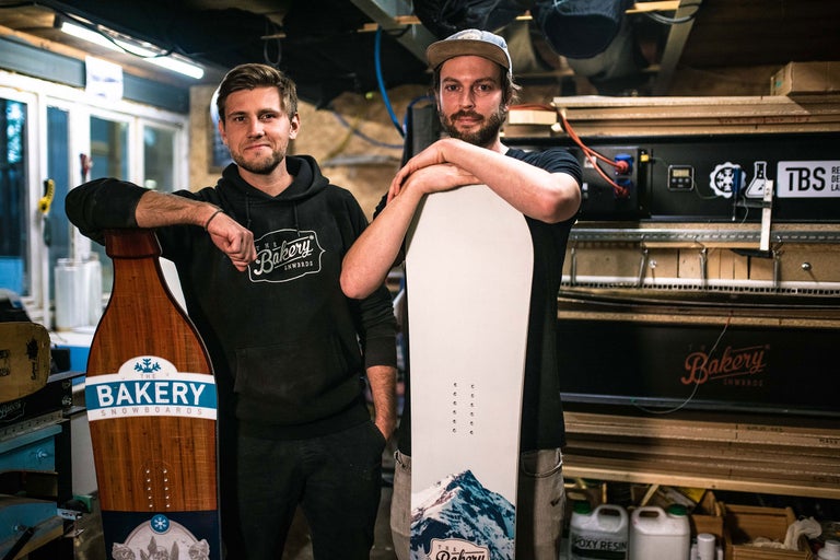 Zwei Personen posieren mit Snowboards der Marke The Bakery Snowboards in einer Werkstatt.