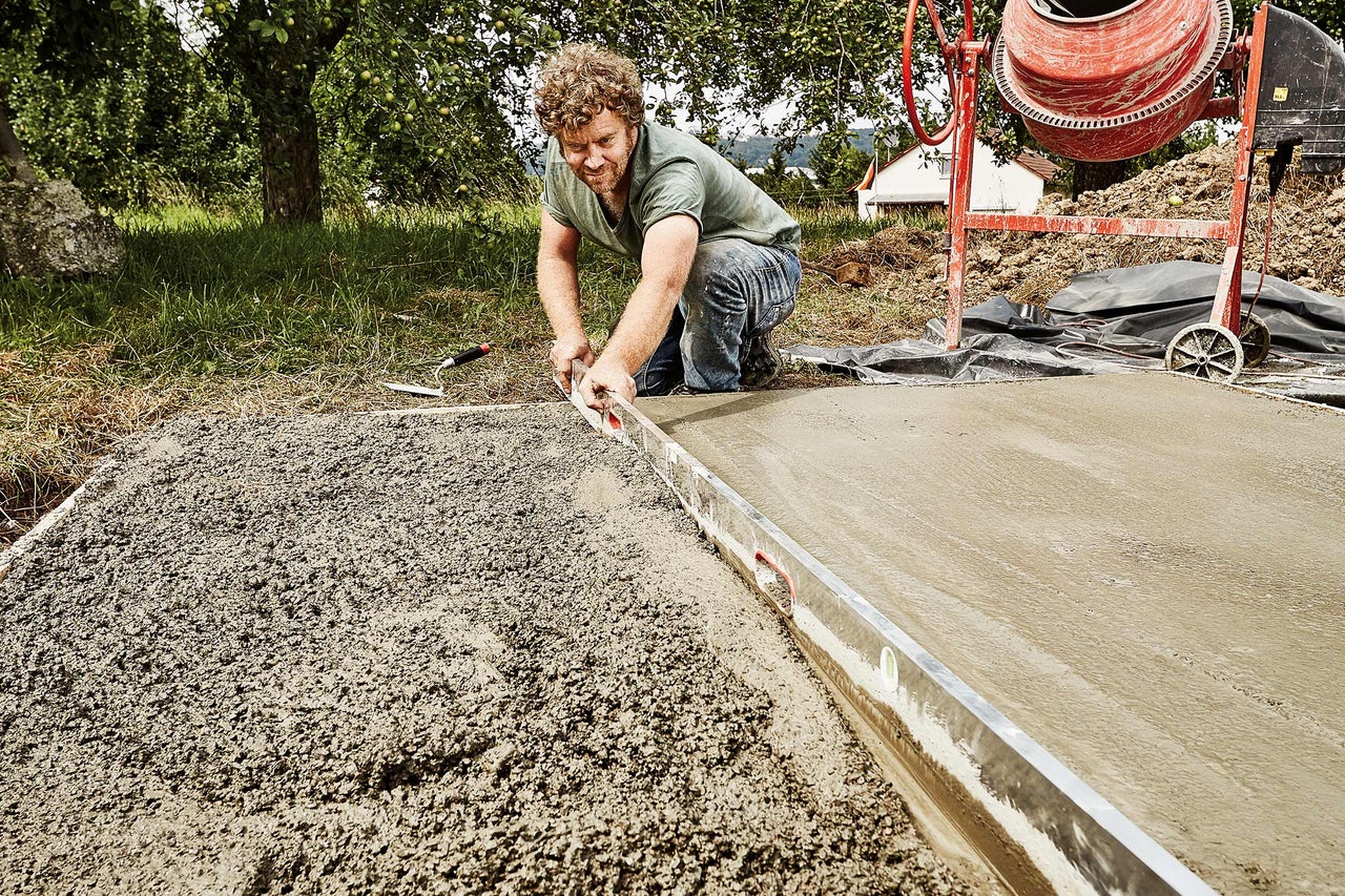 Mann ebnet Beton mit einer Wasserwaage, im Hintergrund ein Betonmischer.