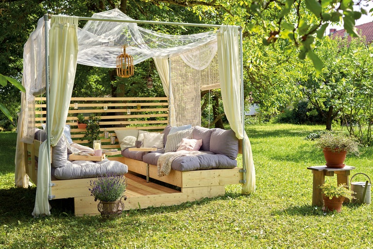 Gartenszene mit überdachter Holzterrasse, Sitzkissen und Pflanzen