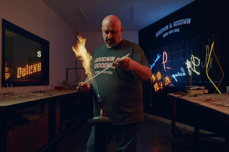 Ein Mann formt in einer Werkstatt ein Neonrohr mit einer Flamme. Im Hintergrund sind gefertigte Neonreklamen zu sehen.