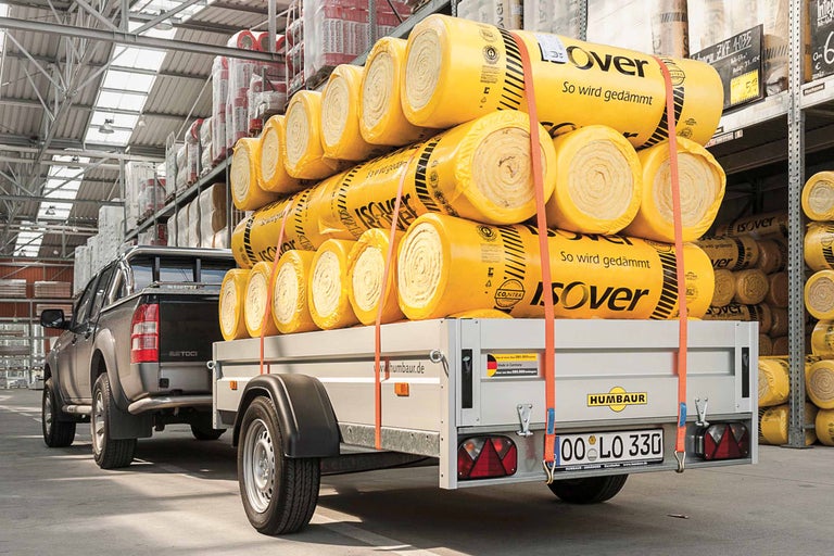 Ein Pick-up zieht einen Anhänger beladen mit Isover Dämmwolle vor einem Regal im Lager.