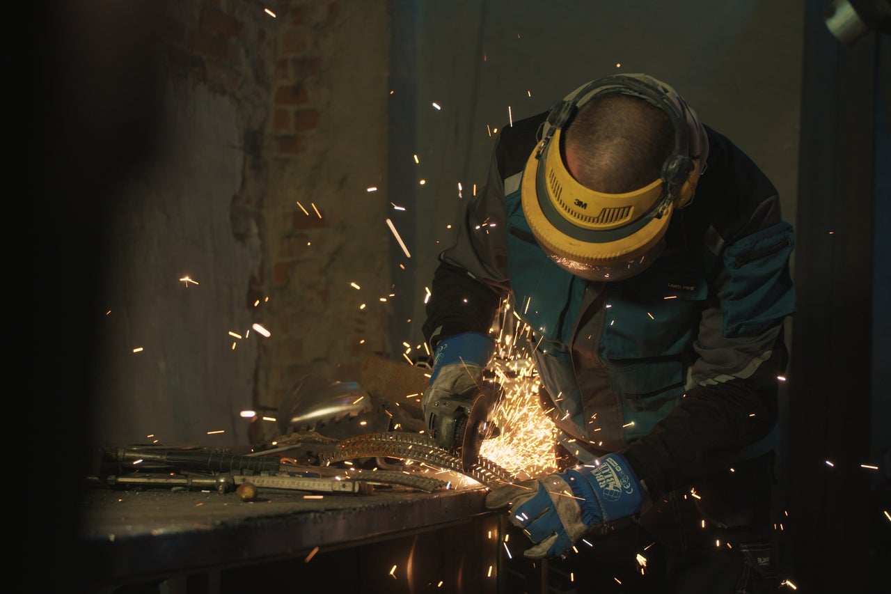 Mann mit Schutzhelm bearbeitet Metall mit einem Winkelschleifer, wobei Funken entstehen.