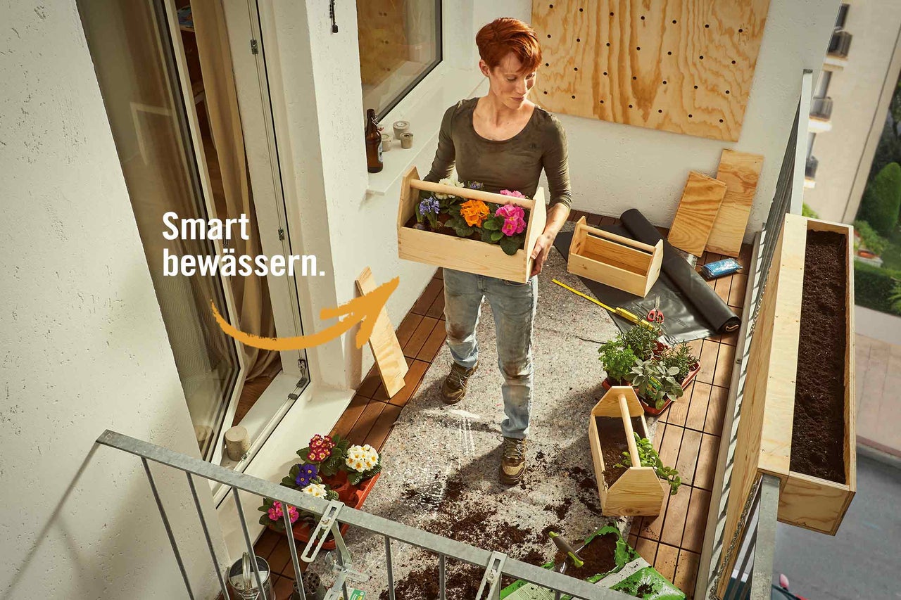 Frau bepflanzt einen Holzkasten mit Blumen auf einem Balkon; im Hintergrund sind weitere Pflanzkästen und Gartenzubehör zu sehen.