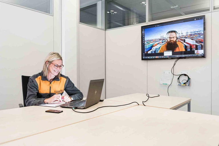 Eine Frau sitzt an einem Tisch und hat ein Videogespräch auf einem Bildschirm an der Wand.
