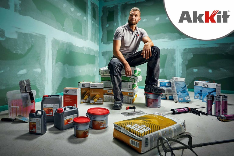 Szene mit verschiedenen Akkit Produkten, darunter Füllstoffe, Spachtelmassen und Klebstoffe, vor einer Baustelle mit verputzten Wänden. Ein Mann sitzt auf einem Stapel Akkit Säcken. Akkit Logo.