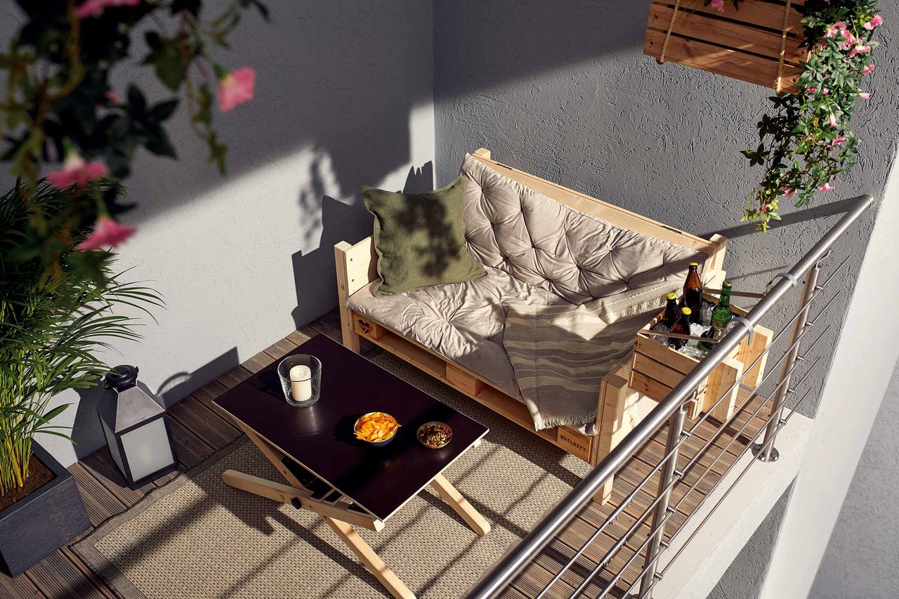 Balkon mit Palettensofa aus Holz, Polstern, Beistelltisch mit Snacks und Kerze, Pflanzen und hölzernem Getränkehalter am Geländer.