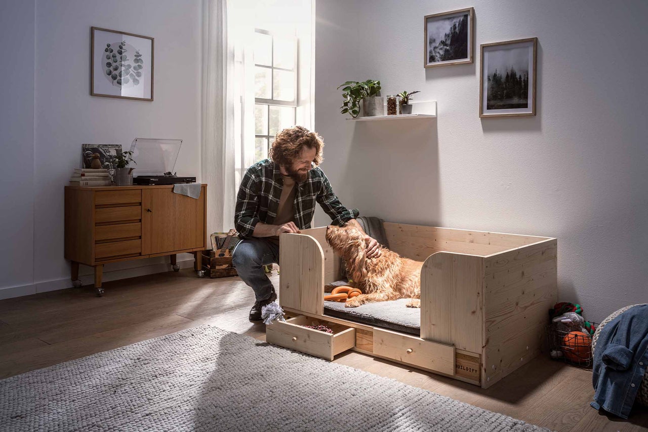 Mann streichelt Hund in einem Hundebett aus Holz mit Schubladen in einem hellen Wohnzimmer.
