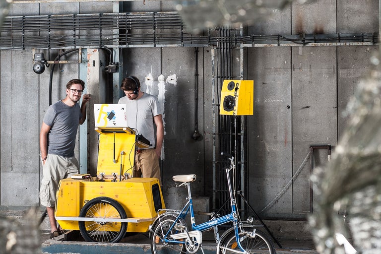 Zwei Personen mit gelbem Transportwagen, Lautsprecher und Fahrrad vor Betonwand.