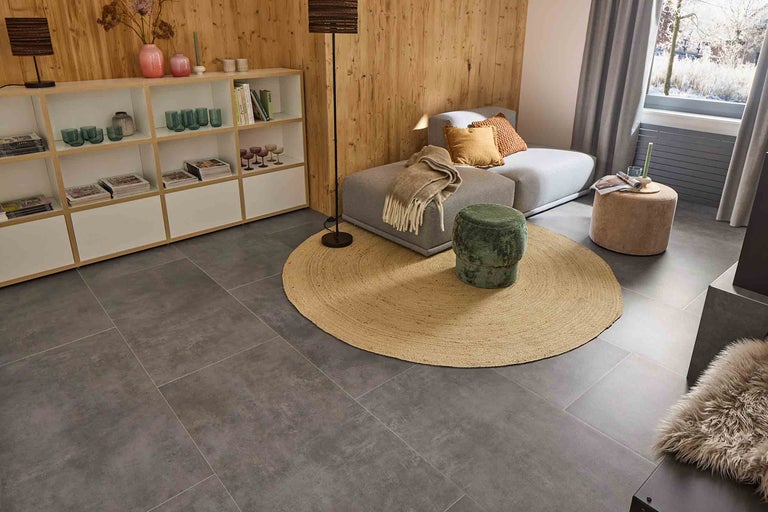 Wohnzimmer mit grauen Bodenfliesen, Sofa, Teppich, Hocker und Regal.