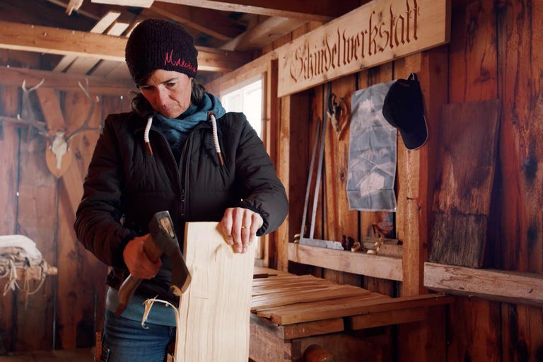 Frau arbeitet mit einem Beil in einer Holzwerkstatt an einem Holzbrett.