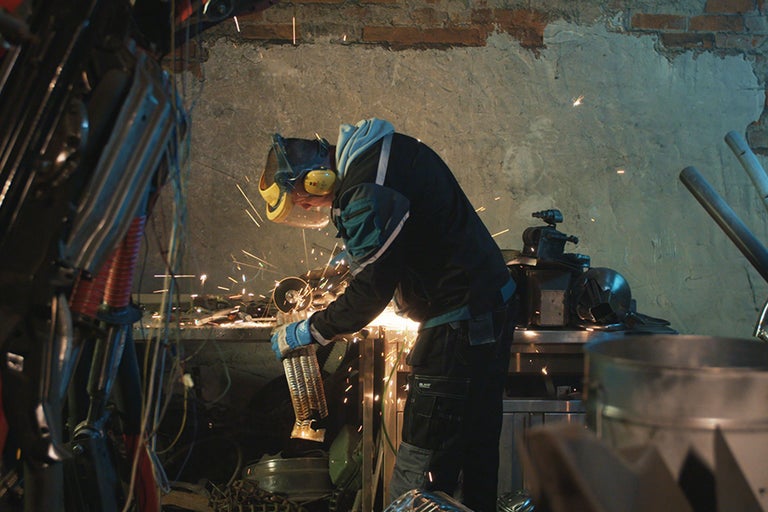 Ein Handwerker mit Schutzmaske bearbeitet ein Metallobjekt mit einer Schleifmaschine in einer Werkstatt.