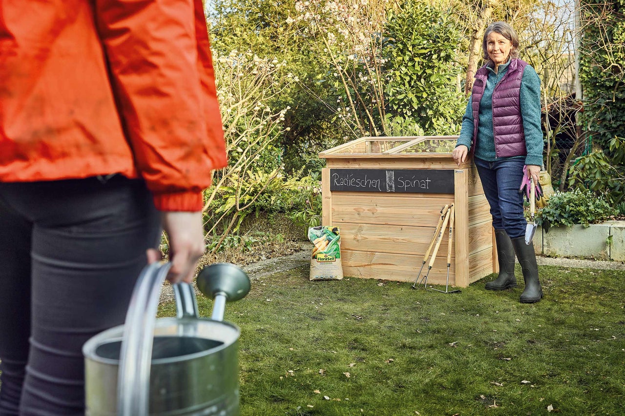 Gartenszene mit Hochbeet, Gießkanne, Frau und Gartenwerkzeug im Garten