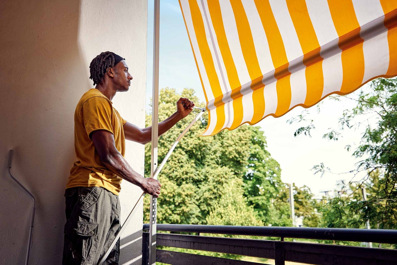 Mann bedient eine gelb gestreifte Markise auf einem Balkon.