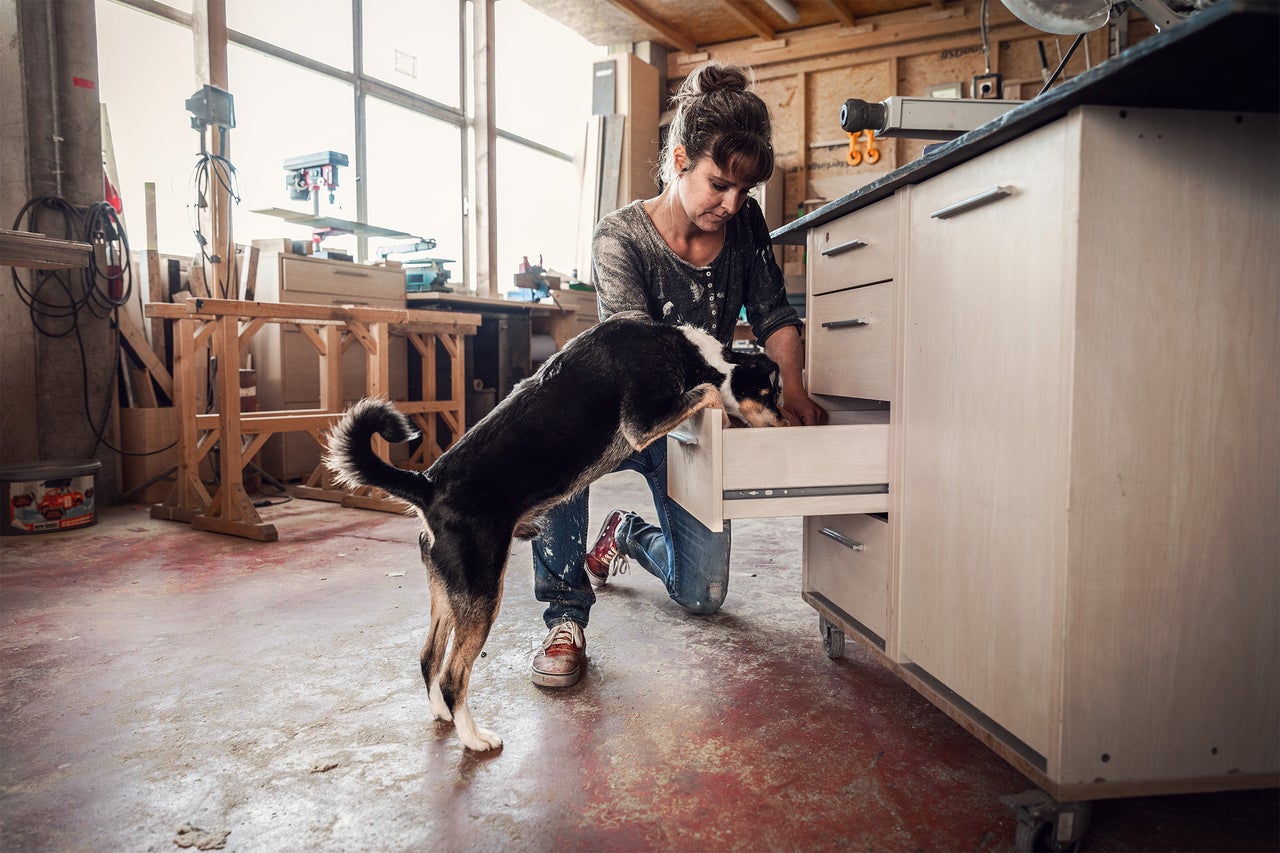 Eine Frau kniet in einer Werkstatt und öffnet eine Schublade, während ein Hund hineinschaut.