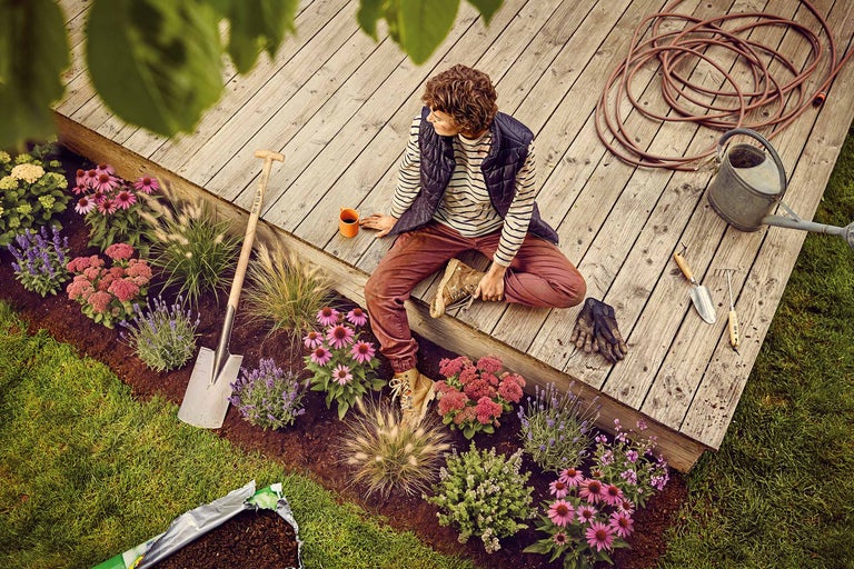 Frau sitzt auf Holzterrasse neben einem bepflanzten Beet mit Spaten, Gießkanne, Gartenschlauch und verschiedenen Blumen wie Sonnenhut und Lavendel.