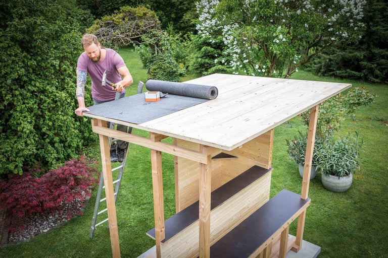 Mann befestigt Dachpappe auf einem Gartenhaus mit Hammer.