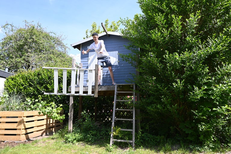 Ein Junge steht auf einem erhöhten Holzspielhaus mit Leiter in einem Garten.