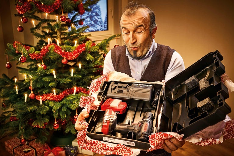 Mann öffnet ein Geschenk mit einem Akkuschrauber Set vor einem Weihnachtsbaum.