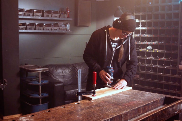 Eine Person arbeitet mit einer Oberfräse an einem Holzstück auf einer Werkbank in einer Werkstatt.