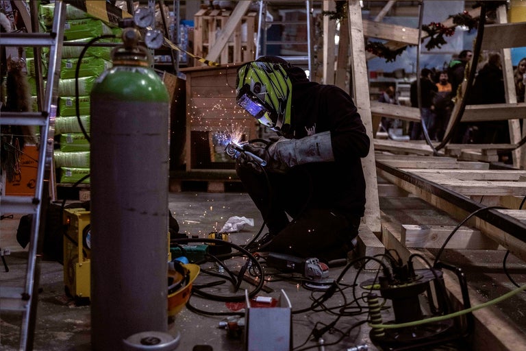 Eine Person mit Schweißhelm schweißt Metall mit einer Schweißmaschine und einer Gasflasche.