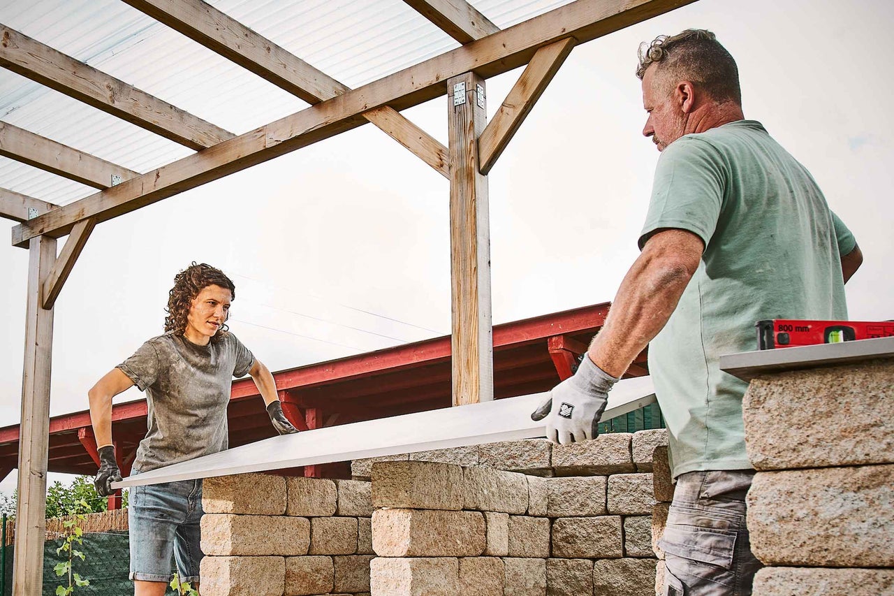 Zwei Personen errichten eine Gartenmauer mit Betonsteinen unter einem hölzernen Unterstand.