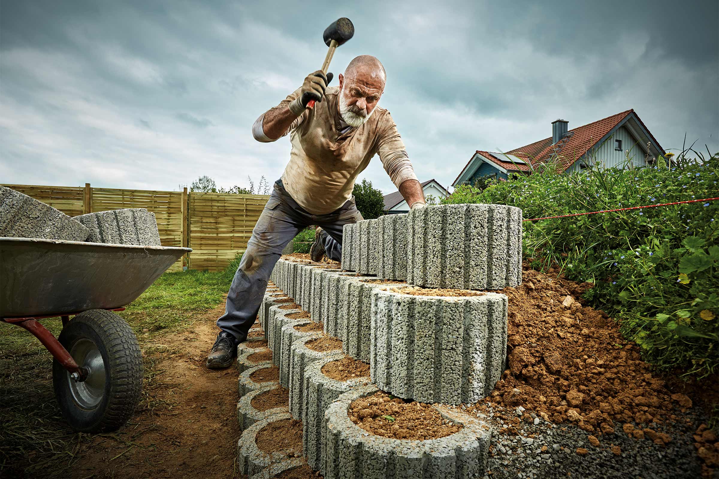 Ein Mann baut mit einem Gummihammer eine Gartenmauer aus Betonsteinen, Schubkarre im Hintergrund.
