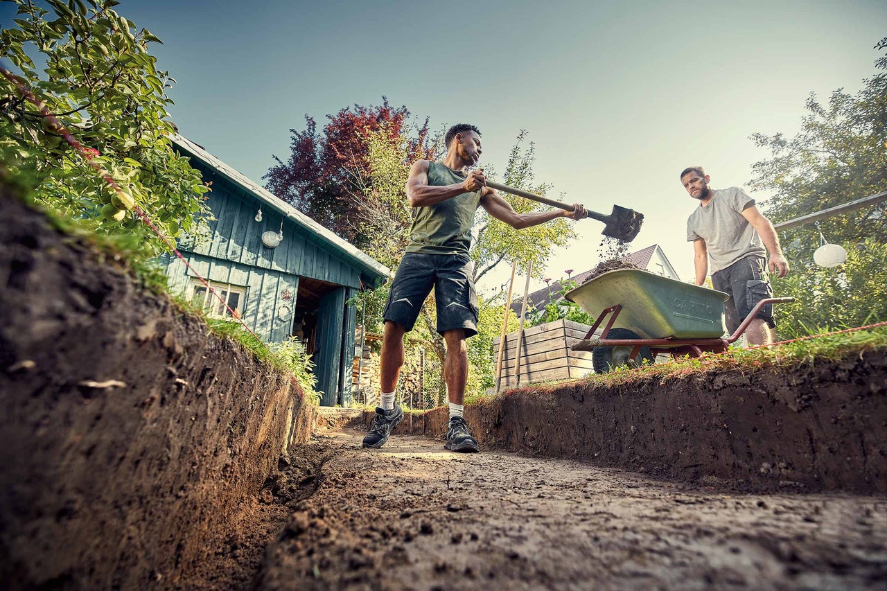 Zwei Männer arbeiten im Garten mit einer Schaufel und einer Schubkarre.