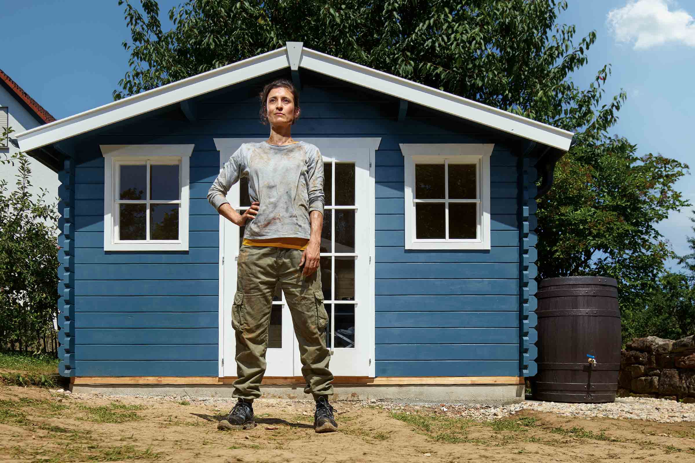 Eine Frau steht vor einem blauen Gartenhaus mit weißen Akzenten, neben dem sich eine Regentonne befindet.