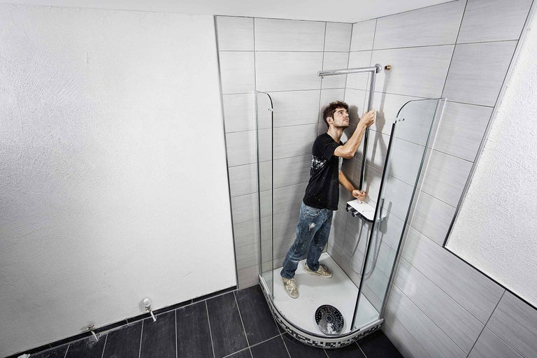 Ein Mann installiert eine Duschkabine in einem Badezimmer mit grauen Fliesen und dunklem Boden.