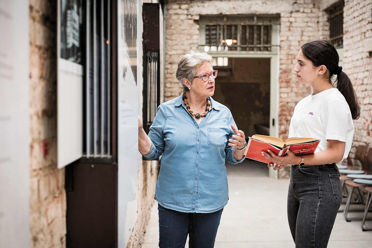 Zwei Frauen diskutieren vor einer Backsteinmauer mit einem Buch in der Hand.