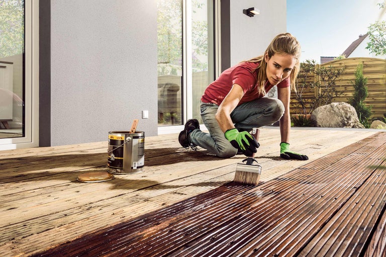 Frau streicht eine Holzterrasse mit einem Pinsel und einer Dose Holzschutzlasur.