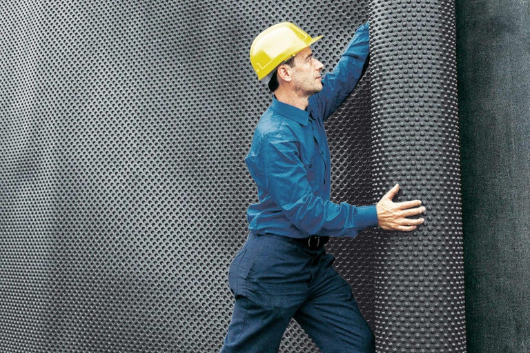 Ein Mann mit Schutzhelm verlegt Noppenbahn zur Bauwerksabdichtung.