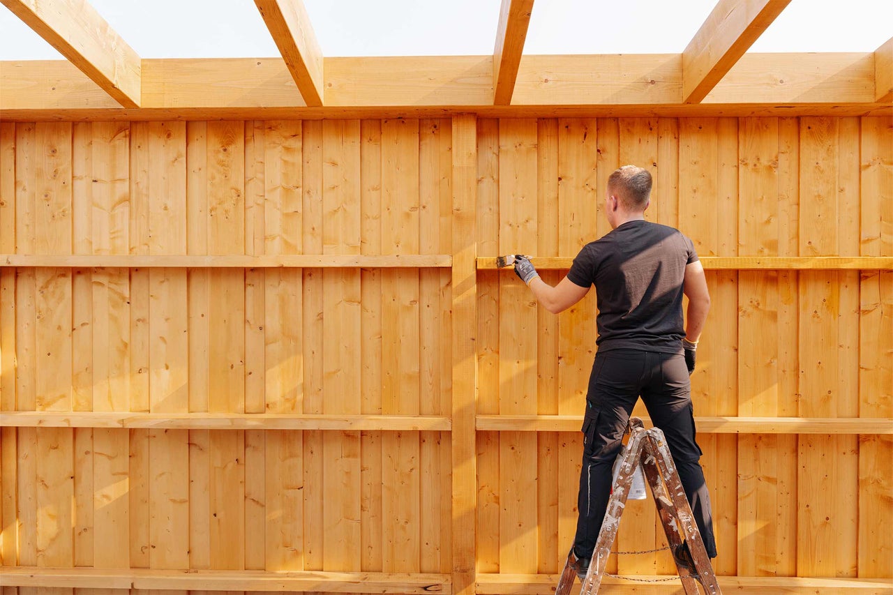 Mann streicht eine Holzkonstruktion mit einem Pinsel von einer Leiter aus.