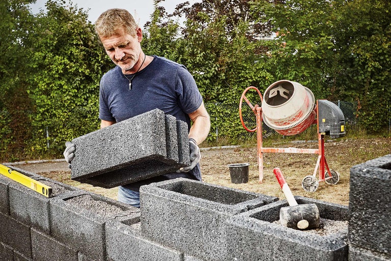 Mann baut eine Mauer mit Mauersteinen, im Hintergrund ein Betonmischer