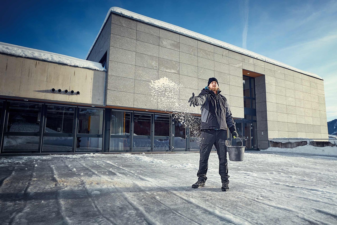 Mann streut Streusalz vor einem Gebäude auf eisigem Untergrund