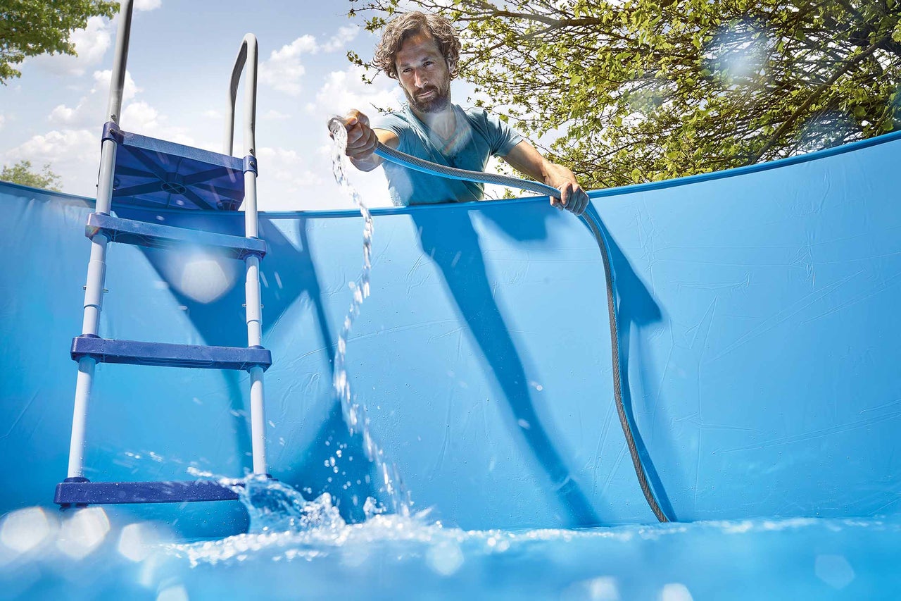 Mann befüllt Schwimmbecken mit Wasserschlauch; Schwimmbadleiter im Bild.