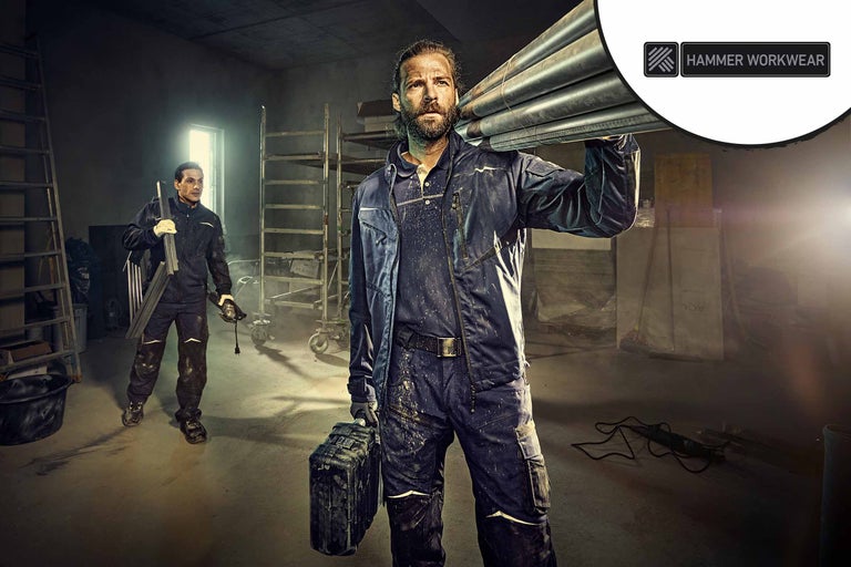 Zwei Arbeiter in Arbeitskleidung in einem hellen, organisierten Raum. Einer trägt Rohre, der andere eine Schleifmaschine. Hammer Workwear Logo