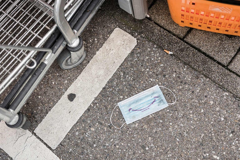 Detailaufnahme eines Einkaufswagens, einer Gesichtsmaske und eines orangefarbenen Korbs auf einer Pflasterfläche.