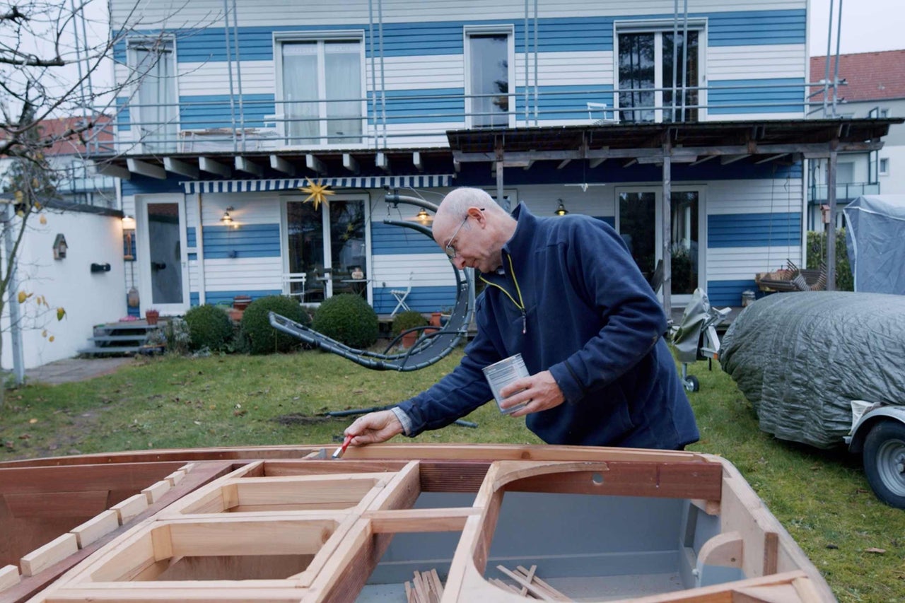Ein Mann streicht ein Holzboot mit einem Pinsel und einer Dose Farbe im Garten eines Hauses.