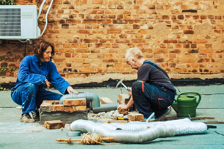 Zwei Personen arbeiten an einer Installation im Freien mit Rohren und Steinen vor einer Ziegelwand.