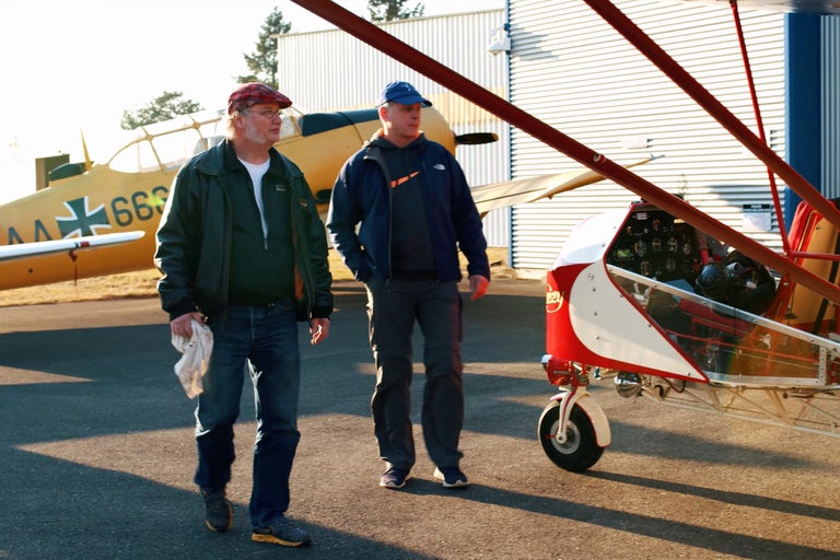 Zwei Männer stehen neben einem Ultraleichtflugzeug auf einem Flugplatz.