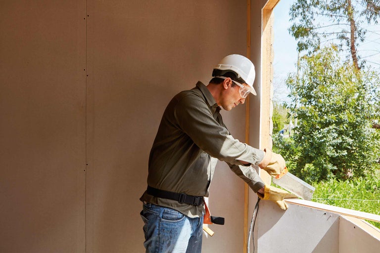 Ein Bauarbeiter mit Schutzhelm und Schutzbrille arbeitet an einem Fensterrahmen auf einer Baustelle.