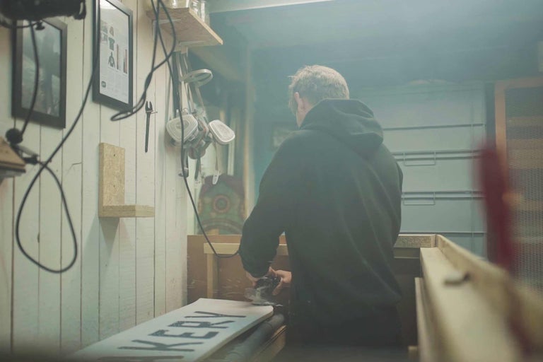 Eine Werkstattszene mit einer Person, die ein Holzbrett bearbeitet. Werkzeuge und Regale sind an den Wänden angebracht.